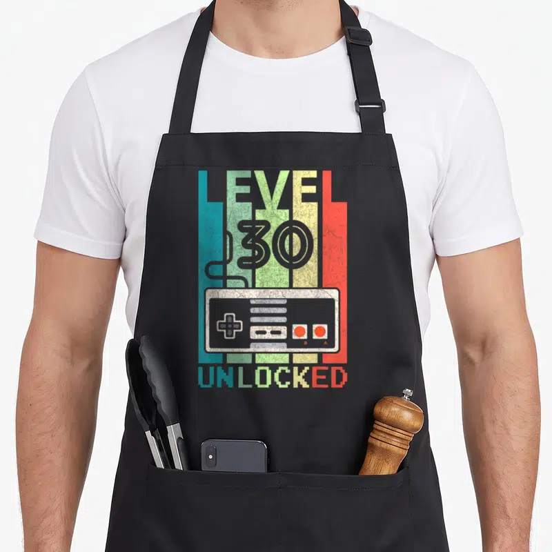 Video gamer 30th birthday apron - Retro game controller with LEVEL 30 UNLOCKED text on four colored vertical stripes — shown on a black cotton canvas apron against a white background