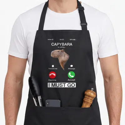 Call animal apron - Capybara incoming call screen with I MUST GO banner and red decline button — shown on a black cotton canvas apron against a white background