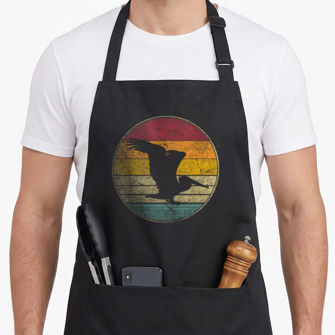 80s bird apron - Grey pelican silhouette over red orange yellow green grey retro stripes — shown on a black cotton canvas apron against a white background