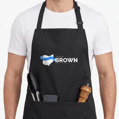 Sherrod briard apron - White Ohio map with blue ribbon reading Blue Wave 2018 next to Sherrod Brown Ohio Senate text — shown on a black cotton canvas apron against a white background