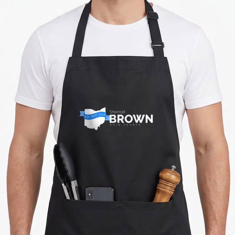Sherrod briard apron - White Ohio map with blue ribbon reading Blue Wave 2018 next to Sherrod Brown Ohio Senate text — shown on a black cotton canvas apron against a white background