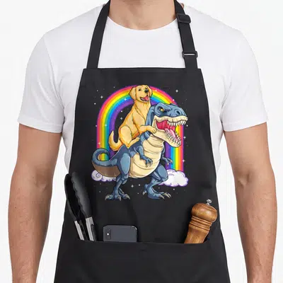 Riding golden retriever apron - Yellow dog riding blue T-Rex under rainbow with stars cartoon — shown on a black cotton canvas apron against a white background