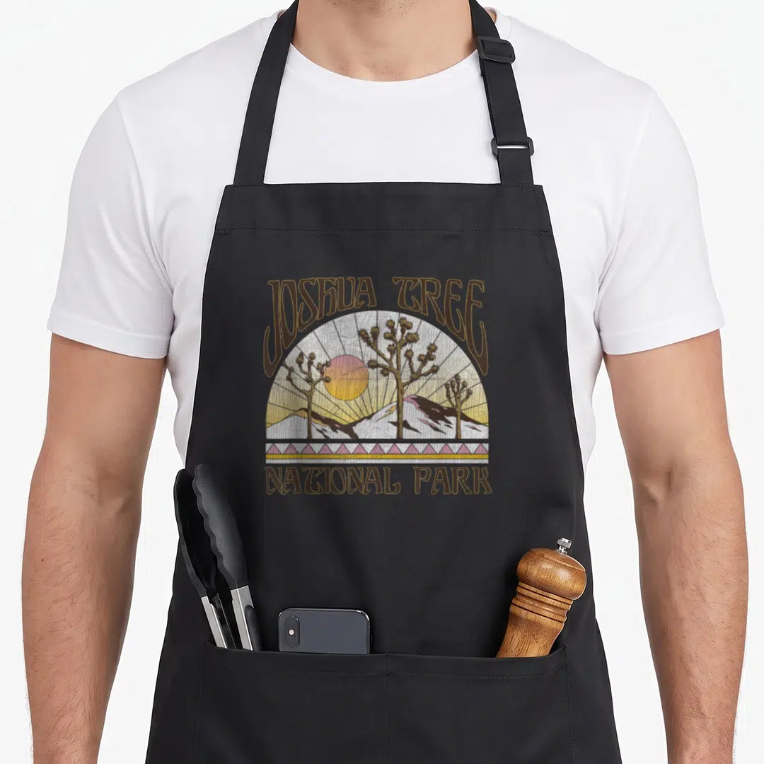 Joshua tree apron - Retro circular desert landscape with Joshua trees, sun, and mountains in brown and warm tones — shown on a black cotton canvas apron against a white background