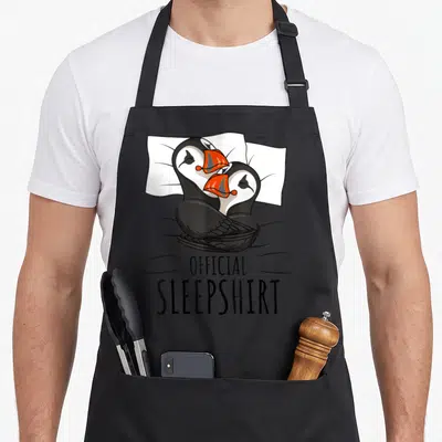 Sleepshirt funny puffin bird apron - Two penguins cuddling under blanket with official sleepshirt text — shown on a black cotton canvas apron against a white background