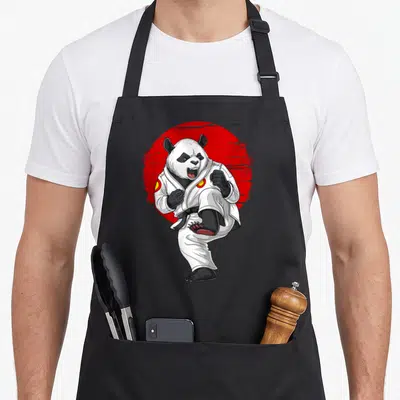 Karate bear apron - Cartoon panda in white karate gi kicking in front of a red distressed circle — shown on a black cotton canvas apron against a white background