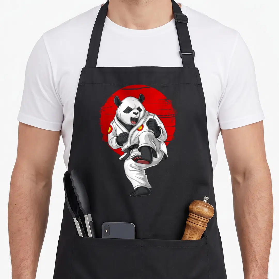 Karate bear apron - Cartoon panda in white karate gi kicking in front of a red distressed circle — shown on a black cotton canvas apron against a white background