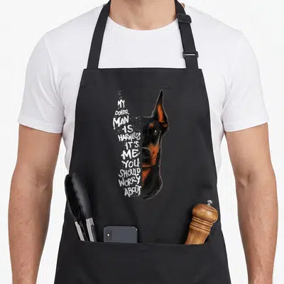 Harmless doberman apron - Stacked white text forming a face shape beside a realistic black and tan Doberman head — shown on a black cotton canvas apron against a white background