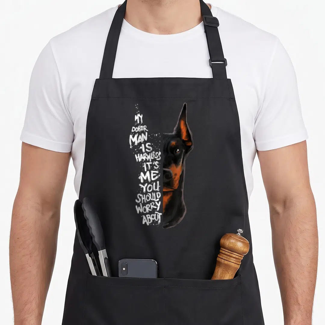 Harmless doberman apron - Stacked white text forming a face shape beside a realistic black and tan Doberman head — shown on a black cotton canvas apron against a white background