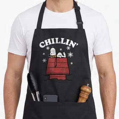 Chillin snow white apron - Arched CHILLIN' text above white dog lying on red doghouse with snowflakes — shown on a black cotton canvas apron against a white background