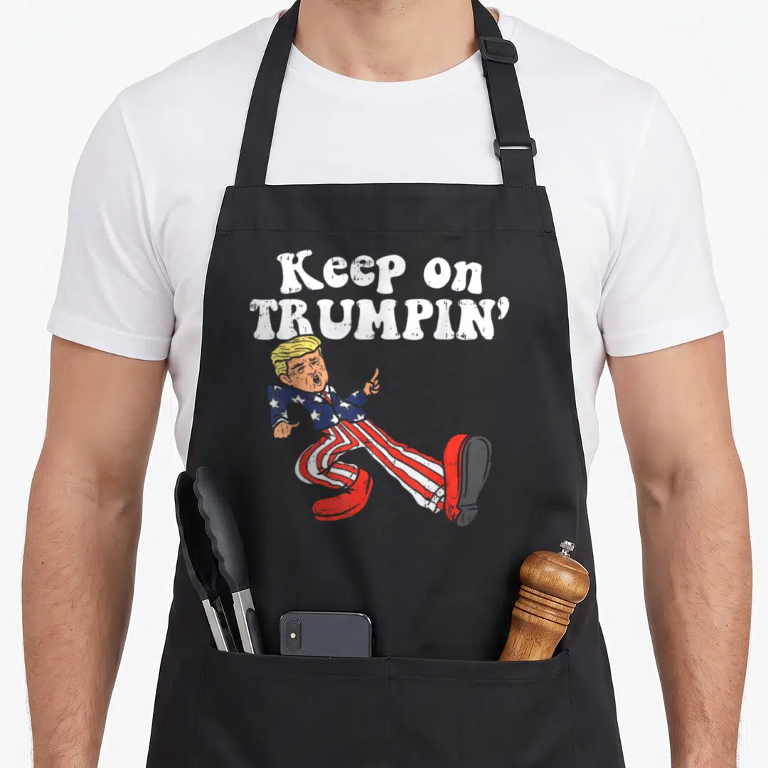 Trumpin funny butter apron - Stacked text Keep on Trumpin above a dancing cartoon figure in a flag suit — shown on a black cotton canvas apron against a white background