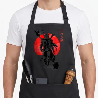 Sun novelty apron - Mandalorian and Grogu silhouette walking in front of a large red circle with vertical Japanese text — shown on a black cotton canvas apron against a white background