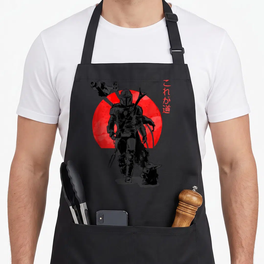 Sun novelty apron - Mandalorian and Grogu silhouette walking in front of a large red circle with vertical Japanese text — shown on a black cotton canvas apron against a white background