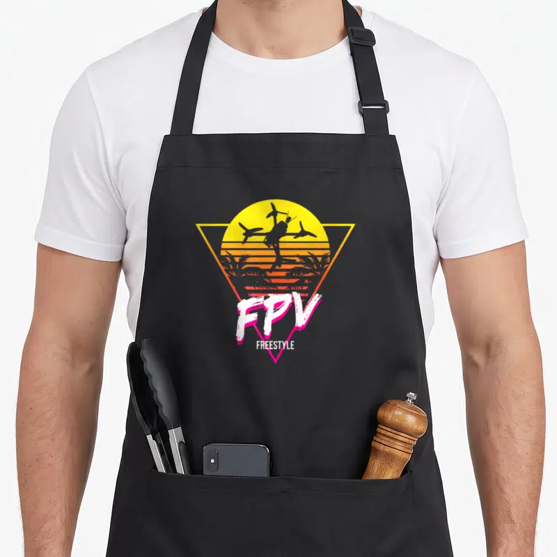 Fpv racing apron - Black drone silhouette inside yellow triangle with striped sunset and FPV FREESTYLE text — shown on a black cotton canvas apron against a white background