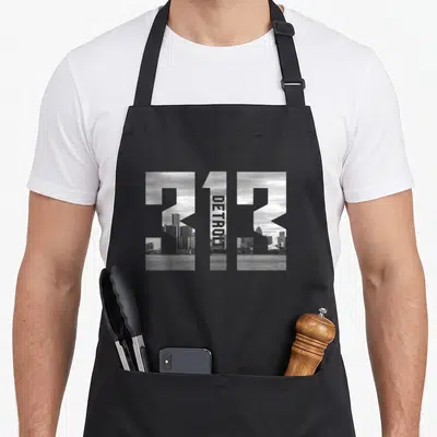 Area detroit apron - 313 numbers filled with grayscale Detroit skyline photo and vertical DETROIT text inside the one — shown on a black cotton canvas apron against a white background