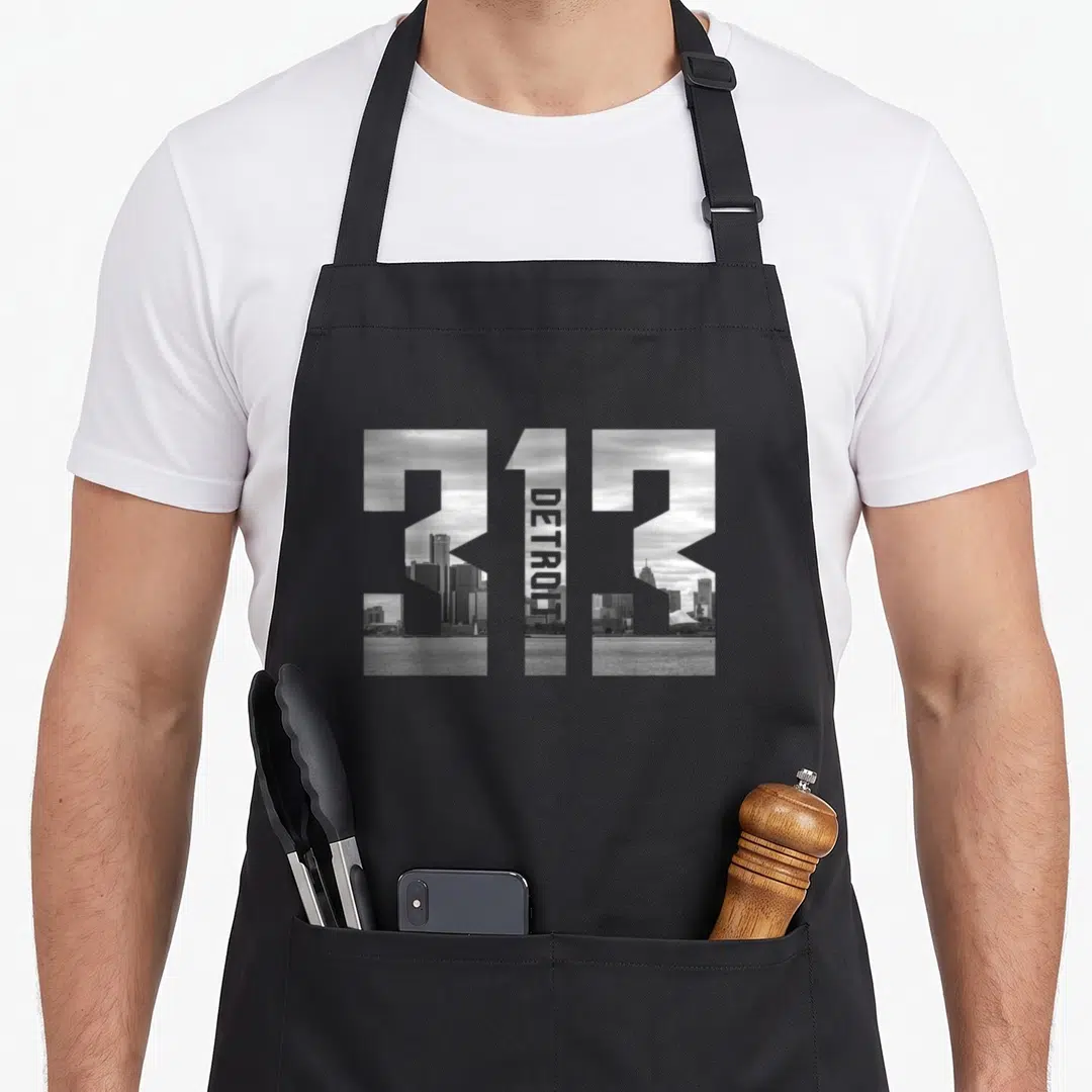 Area detroit apron - 313 numbers filled with grayscale Detroit skyline photo and vertical DETROIT text inside the one — shown on a black cotton canvas apron against a white background