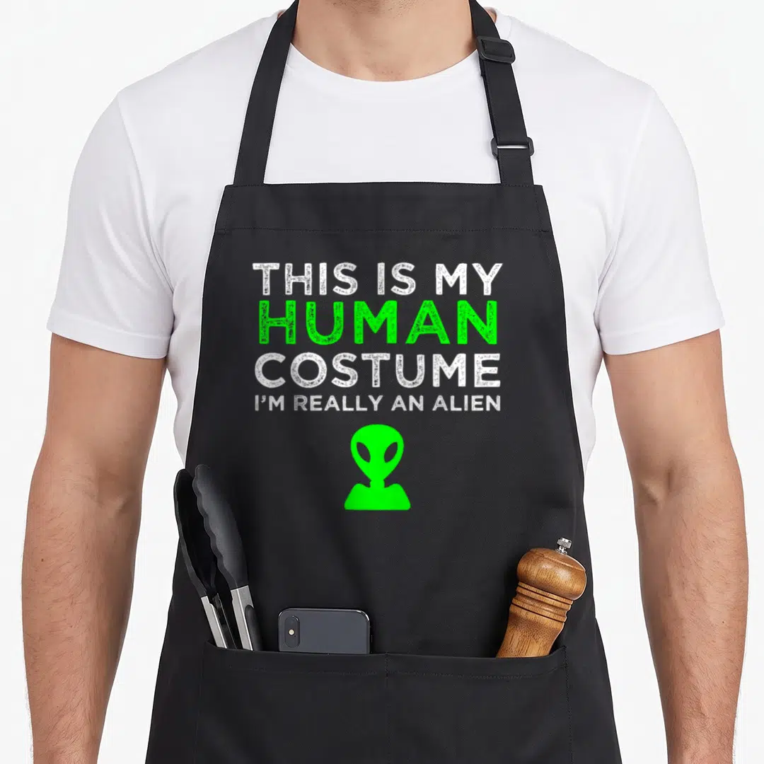 Human funny alien apron - White and green text reading THIS IS MY HUMAN COSTUME above green alien head silhouette — shown on a black cotton canvas apron against a white background