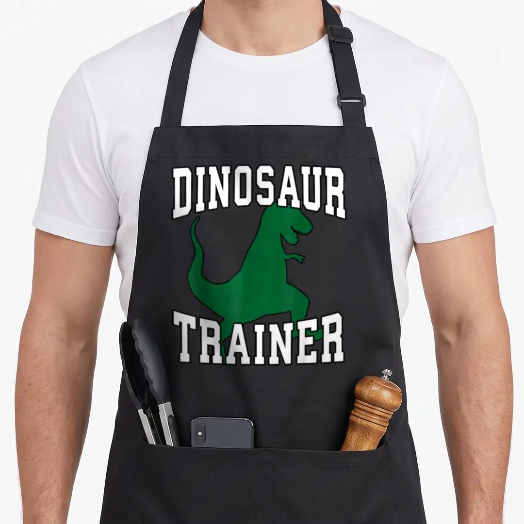 Dinosaur halloween white apron - Green cartoon dinosaur between arched white text DINOSAUR TRAINER — shown on a black cotton canvas apron against a white background