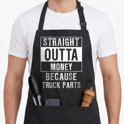 Compton truck apron - Distressed white and grey text reading STRAIGHT OUTTA MONEY BECAUSE TRUCK PARTS stacked vertically — shown on a black cotton canvas apron against a white background