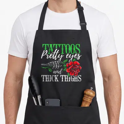 Pretty tattoo apron - Skeleton hand holding red rose with text TATTOOS Pretty eyes and THICK THIGHS — shown on a black cotton canvas apron against a white background