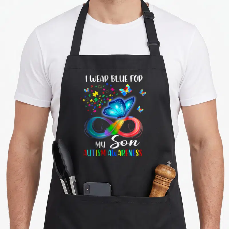 Blue dad and son aprons - I Wear Blue For My Son text with blue butterfly, rainbow infinity symbol, feather, and puzzle — shown on a black cotton canvas apron against a white background