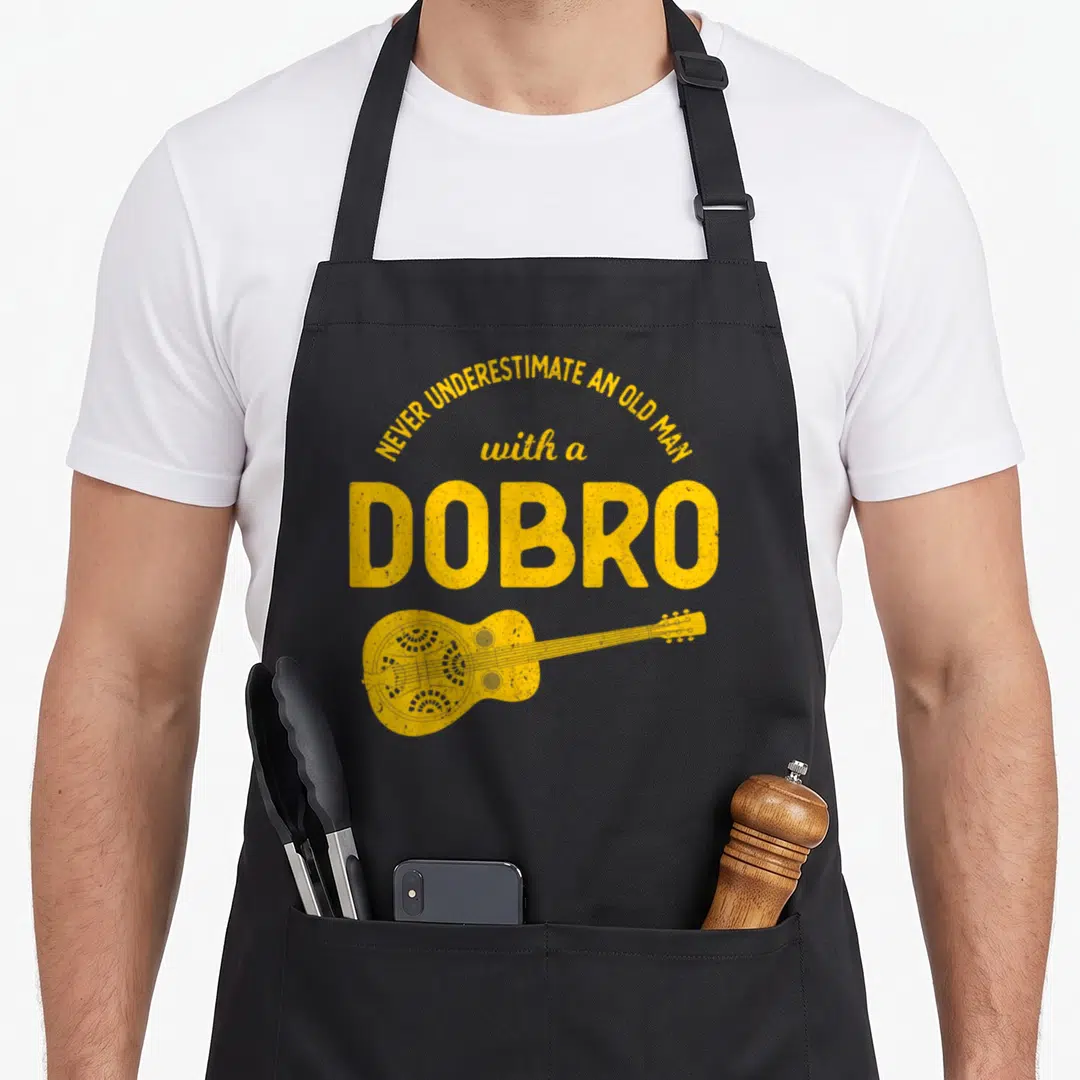 Underestimate bluegrass apron - Yellow retro dobro guitar and stacked text reading NEVER UNDERESTIMATE AN OLD MAN — shown on a black cotton canvas apron against a white background