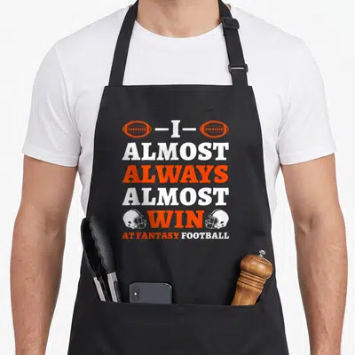 Almost football apron - Stacked orange and white text I Almost Always Almost Win flanked by footballs and helmets — shown on a black cotton canvas apron against a white background