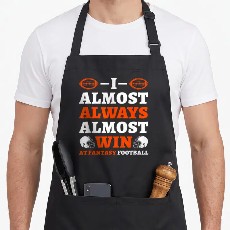 Almost football apron - Stacked orange and white text I Almost Always Almost Win flanked by footballs and helmets — shown on a black cotton canvas apron against a white background