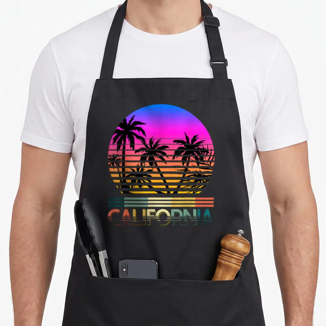Tropical summer aprons - Retro California sunset with sliced palm trees and striped text on canvas — shown on a black cotton canvas apron against a white background