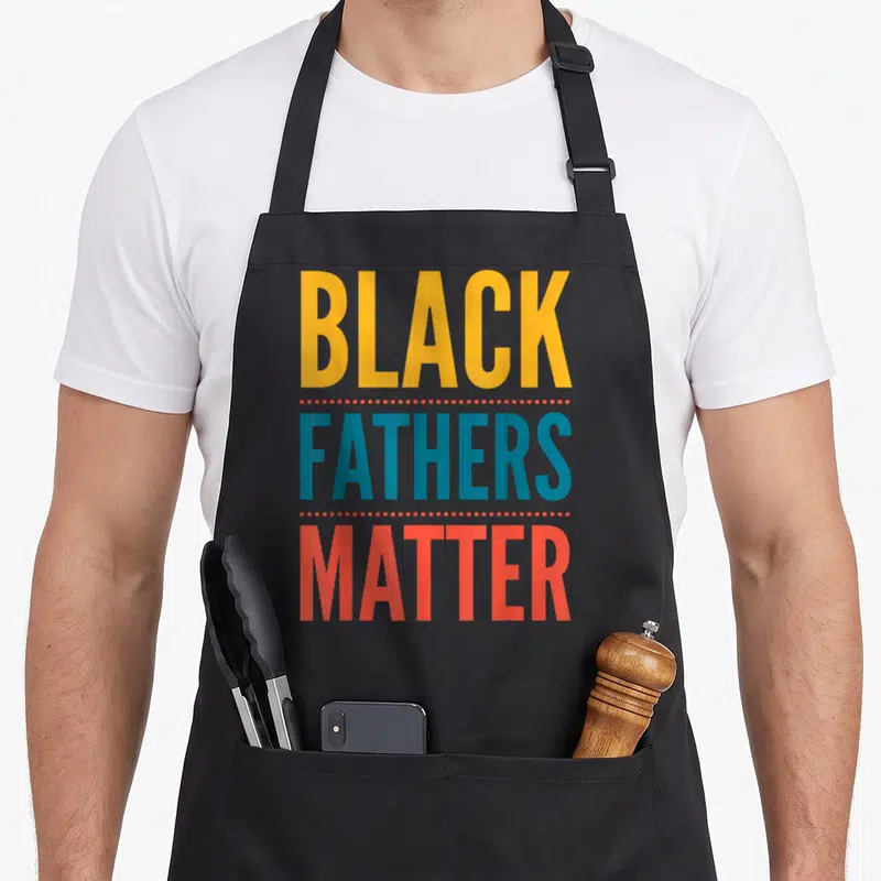 Father business apron - Stacked text reading Black Fathers Matter in yellow blue and red separated by rows of dots — shown on a black cotton canvas apron against a white background