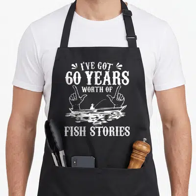 Fisherman 60th birthday apron - Distressed white text sandwiching hands framing a fisherman in a boat on water — shown on a black cotton canvas apron against a white background