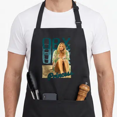 Outer princess apron - Woman sitting on sand with teal OBX text above and Princess script below — shown on a black cotton canvas apron against a white background