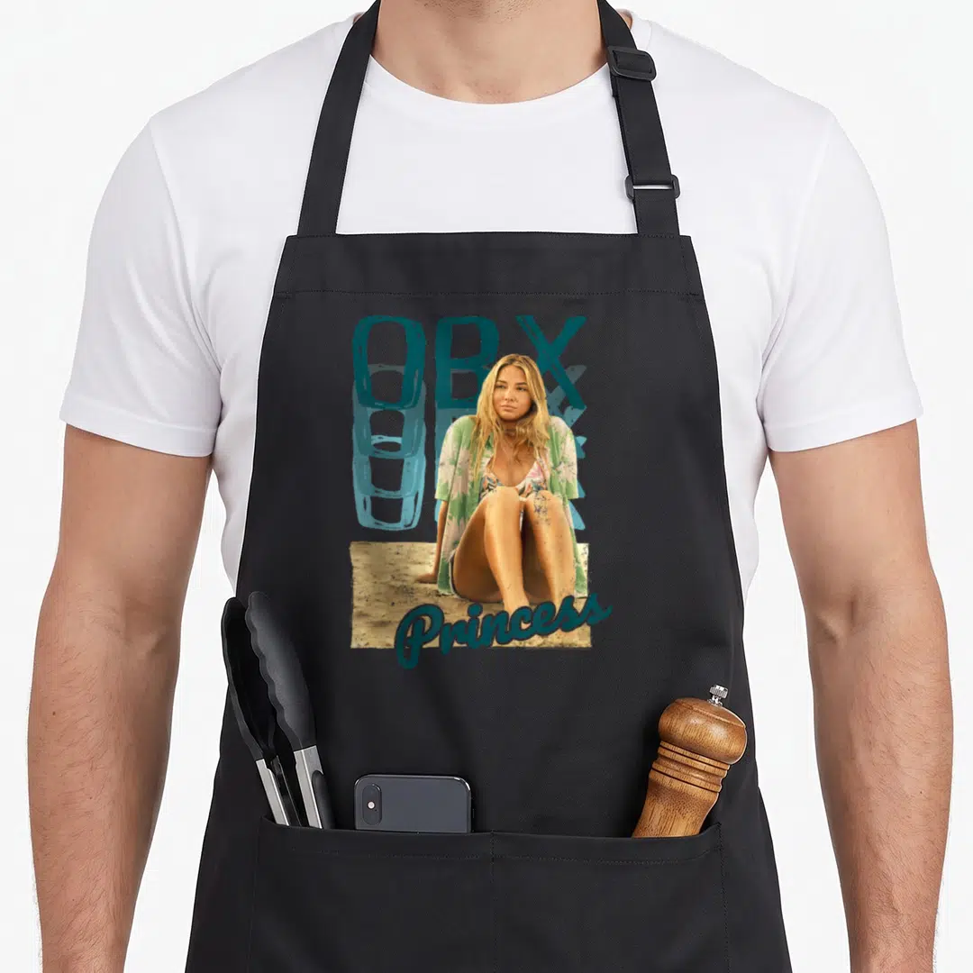 Outer princess apron - Woman sitting on sand with teal OBX text above and Princess script below — shown on a black cotton canvas apron against a white background