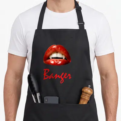 Fang blood apron - Glossy red lips showing white vampire fangs above red cursive text Banger — shown on a black cotton canvas apron against a white background