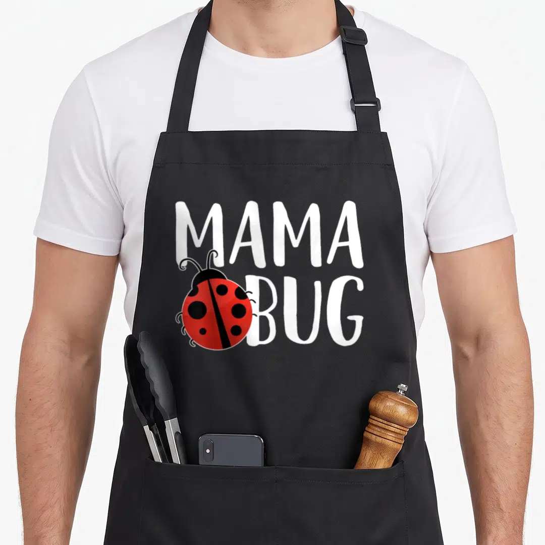 Mama ladybug apron - Cartoon ladybug overlapping white MAMA BUG text in red and black — shown on a black cotton canvas apron against a white background