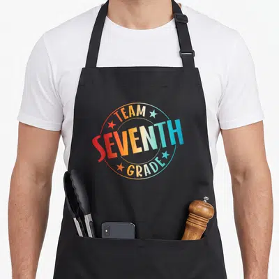 Seventh teacher apron - Team Seventh Grade text in rainbow gradient circle with four stars — shown on a black cotton canvas apron against a white background