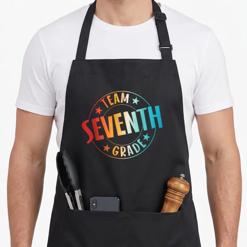 Seventh teacher apron - Team Seventh Grade text in rainbow gradient circle with four stars — shown on a black cotton canvas apron against a white background