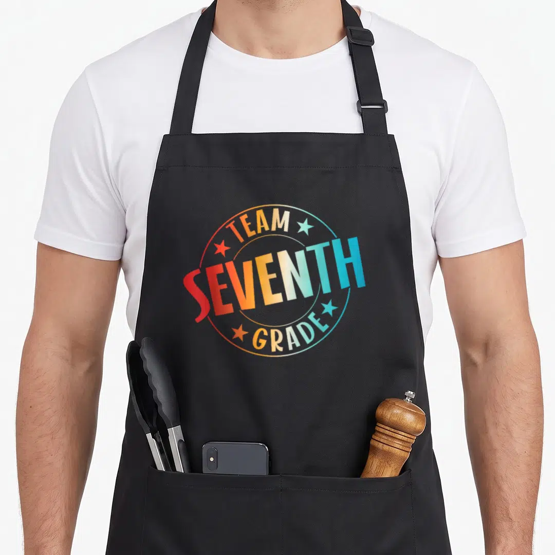 Seventh teacher apron - Team Seventh Grade text in rainbow gradient circle with four stars — shown on a black cotton canvas apron against a white background