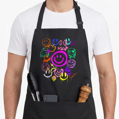 Vivid face on apron - Cluster of dripping cartoon smiley faces in magenta orange blue green purple yellow red — shown on a black cotton canvas apron against a white background