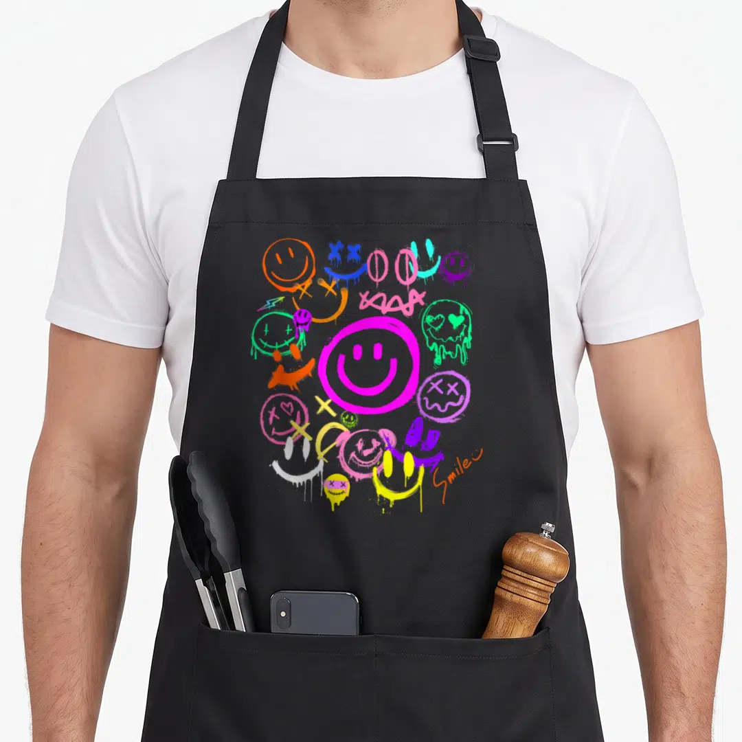 Vivid face on apron - Cluster of dripping cartoon smiley faces in magenta orange blue green purple yellow red — shown on a black cotton canvas apron against a white background