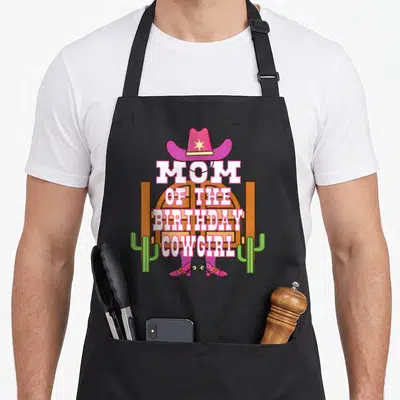 Birthday cowgirl aprons - MOM OF THE BIRTHDAY COWGIRL text with pink hat, boots, and green cacti on brown door — shown on a black cotton canvas apron against a white background