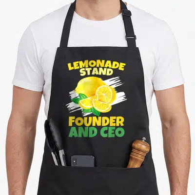 Squad lemonade apron - Realistic yellow lemons and green leaves with founder and ceo text on white brushstroke — shown on a black cotton canvas apron against a white background