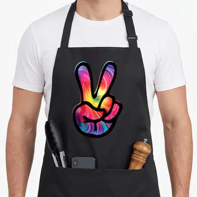 60s tie-dye apron - Cartoon peace sign hand filled with swirling pink, yellow, blue, and purple tie-dye patterns — shown on a black cotton canvas apron against a white background