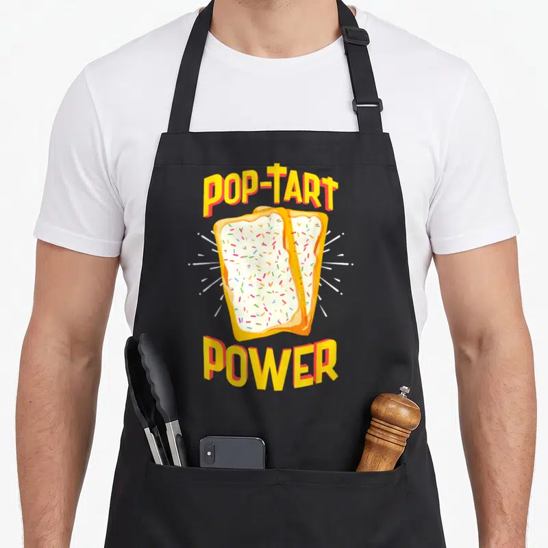 Pop tart apron - POP-TART POWER text above two frosted pastries with white rays in yellow orange white — shown on a black cotton canvas apron against a white background