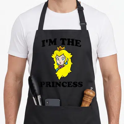Peach shot princess apron - I'M THE PRINCESS text surrounding a cartoon headshot of a blonde princess with a gold crown — shown on a black cotton canvas apron against a white background