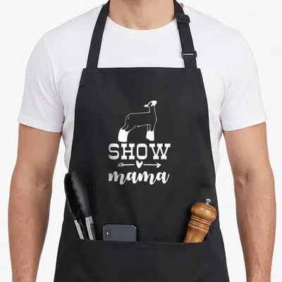 Lamb cooking mama apron - White sheep silhouette above SHOW text and arrow with heart pointing to mama — shown on a black cotton canvas apron against a white background