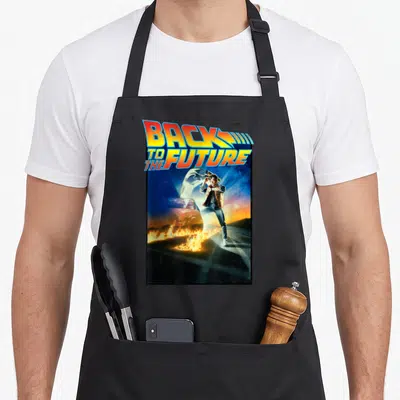 Movie future apron - Back to the Future text over Marty McFly standing on a road with fire trails in yellow orange blue — shown on a black cotton canvas apron against a white background