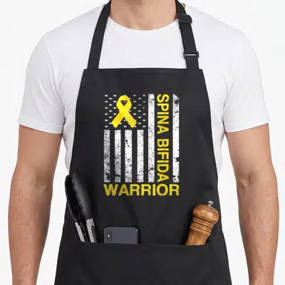 Spina bifida ribbon apron - Yellow ribbon over distressed white flag with vertical SPINA BIFIDA and horizontal WARRIOR — shown on a black cotton canvas apron against a white background