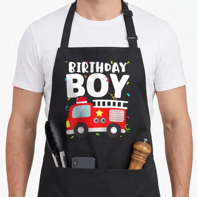 Theme truck apron - Cartoon red fire truck with BIRTHDAY BOY text and colorful confetti scattered around the print — shown on a black cotton canvas apron against a white background