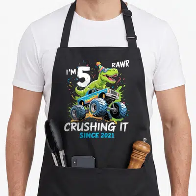 5th birthday monster dinosaur truck apron - Green T-Rex in sunglasses on blue monster truck with party hat and text — shown on a black cotton canvas apron against a white background