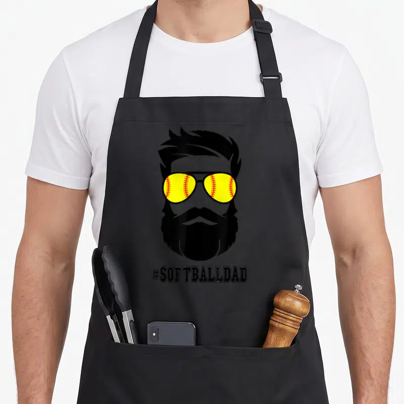 Softball beard apron - Bearded man silhouette with yellow softball sunglasses and #SOFTBALLDAD text — shown on a black cotton canvas apron against a white background